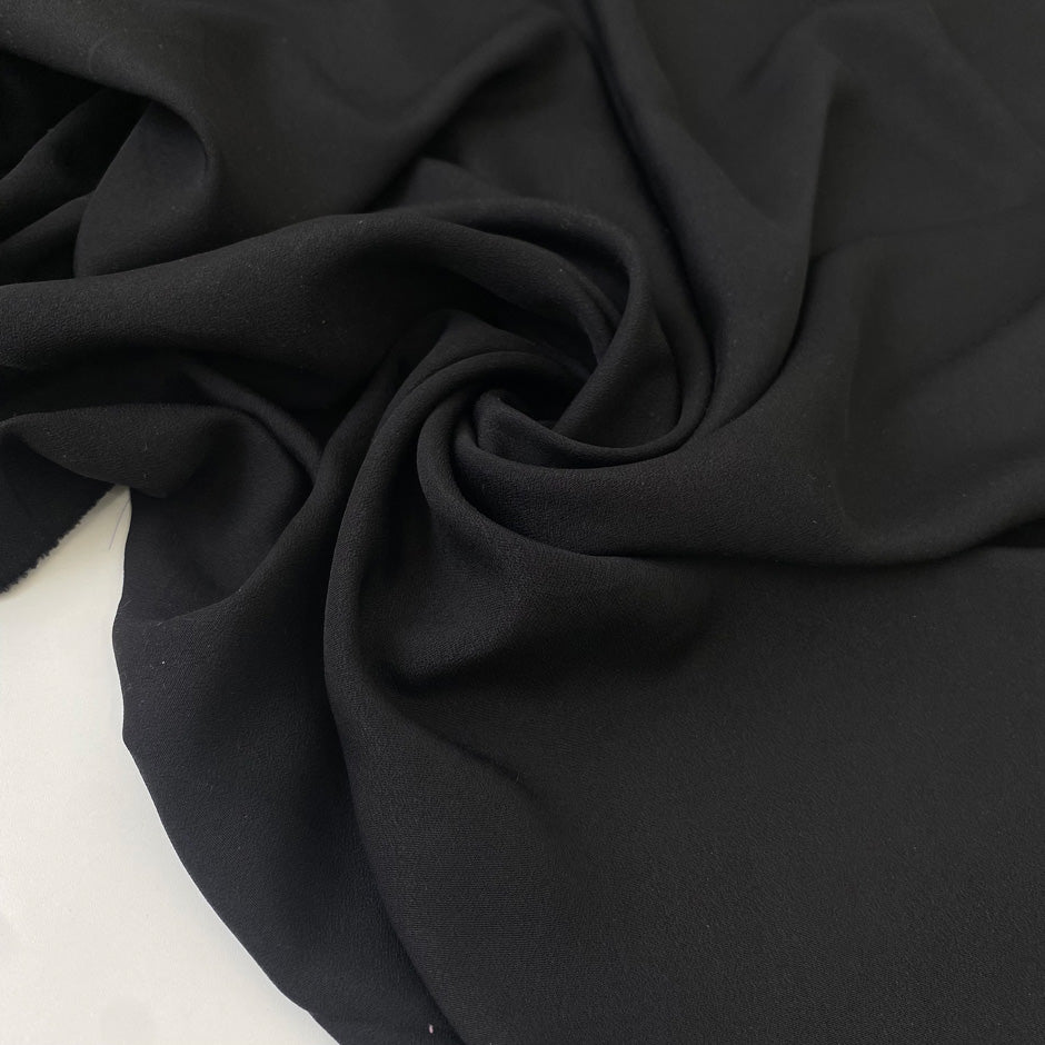 Black polyester crêpe with a fluid drape and slightly textured matte surface ideal for dresses trousers and soft tailoring. High-quality deadstock collected in Stock from a Maison de Couture in Italy.